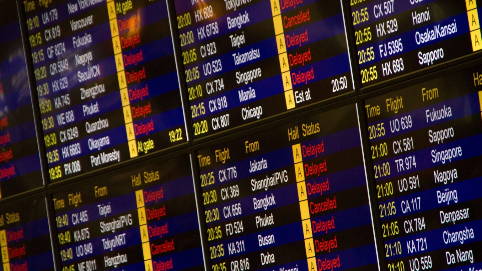 Airport departure board showing flights to cities across Asia with many marked "Delayed" or "Cancelled" in red.
