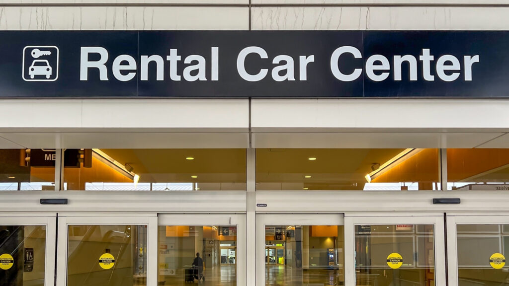 Entrance to a rental car facility with a large overhead sign reading "Rental Car Center." Glass automatic doors lead into the transportation facility.