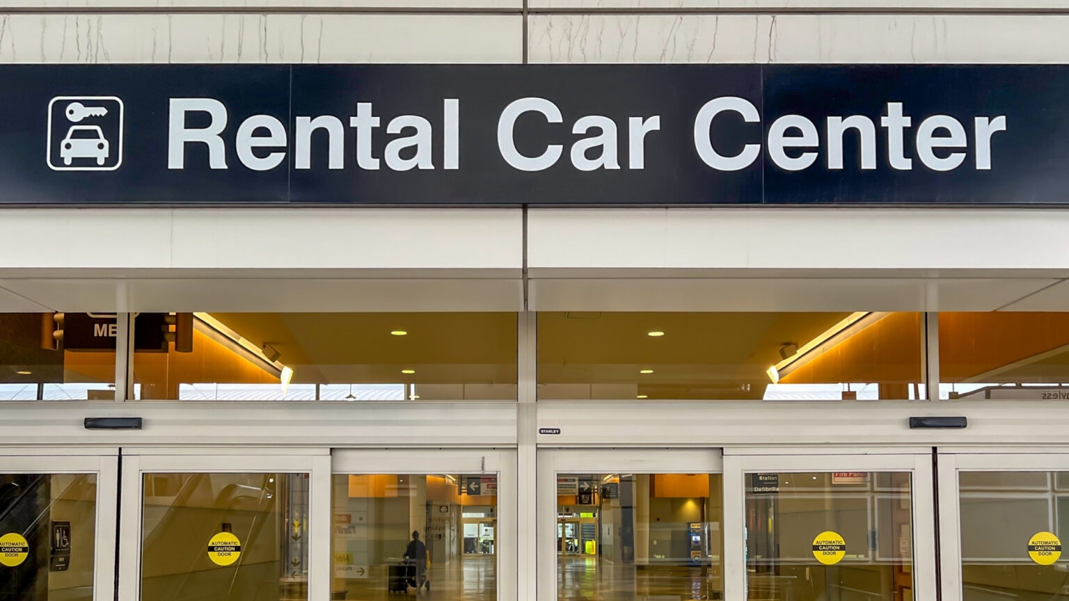 Rental Car Experts Say This One Photo Could Protect You From Extra Charges Entrance to a rental car facility with a large overhead sign reading "Rental Car Center." Glass automatic doors lead into the transportation facility.