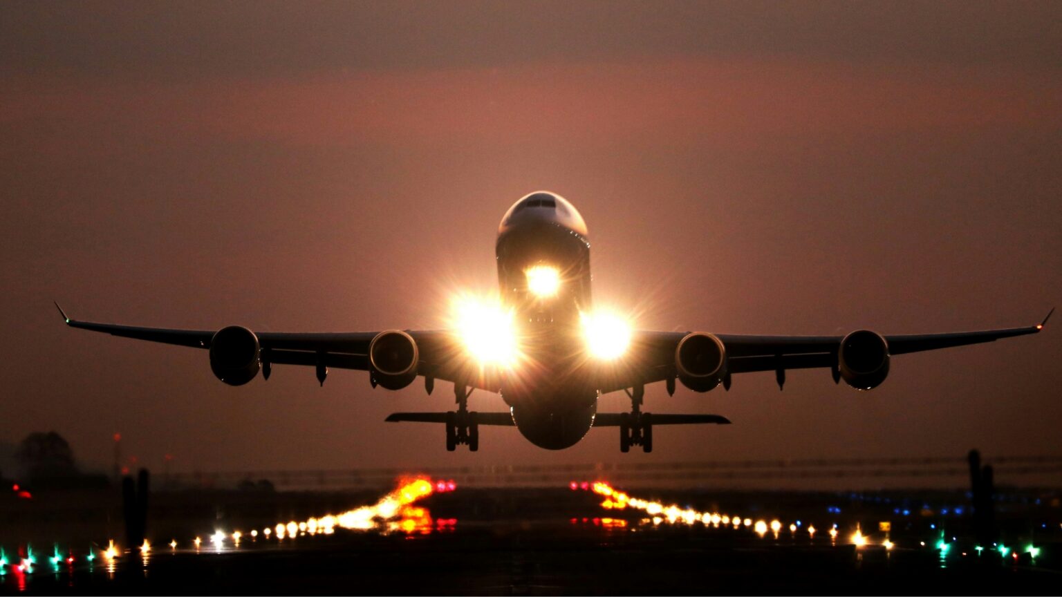 Flying From These U.S. Airports Could Make It Easier to Score a Cheap Flight Large passenger airplane approaches a runway at dusk with its landing gear down and bright landing lights shining toward the camera.