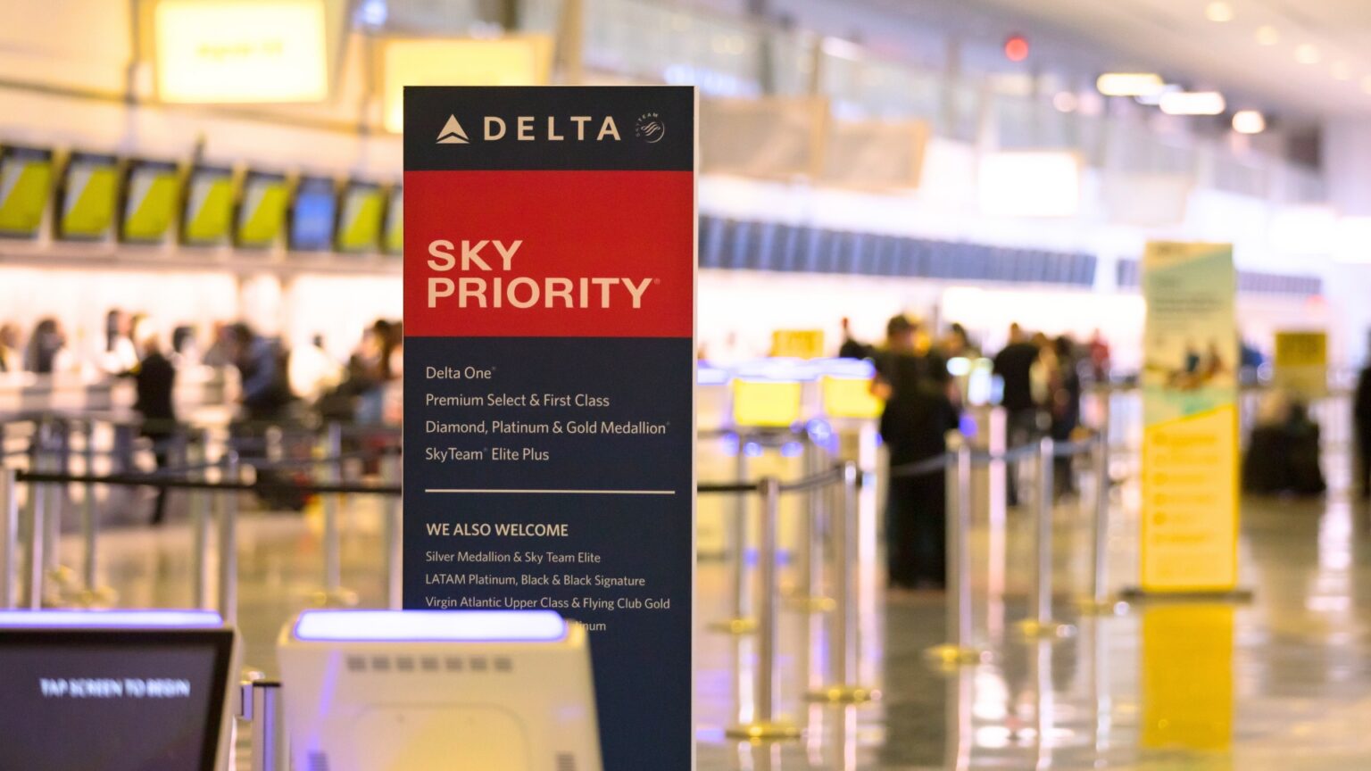 Delta Sky Priority sign stands in an airport check in area with passengers and service counters blurred in the background. The sign lists priority access for Delta One. Premium Select and First Class. Diamond. Platinum and Gold Medallion. SkyTeam Elite Plus.