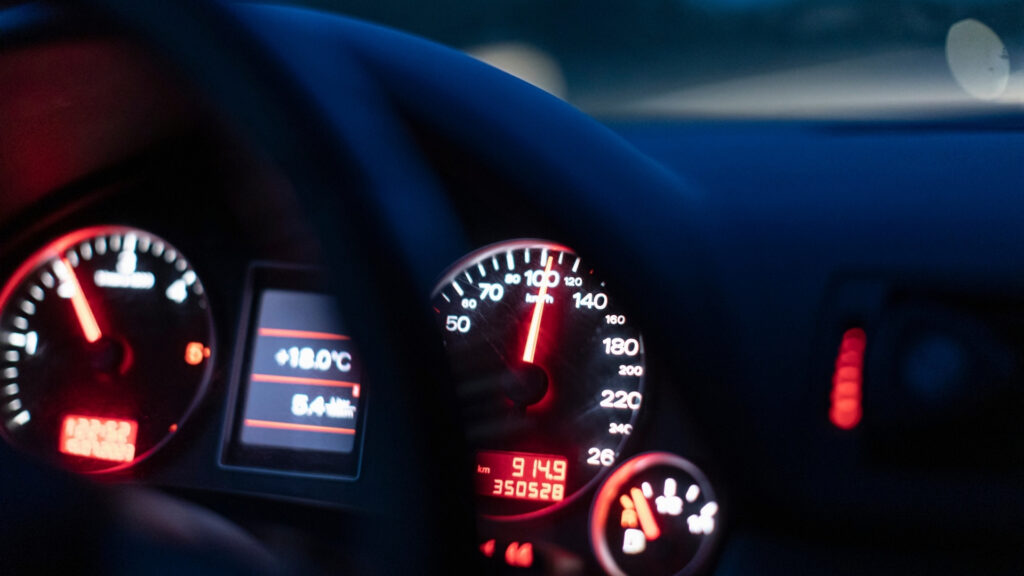 Illuminated car dashboard at night with the speedometer near 100 kilometers per hour. The display reads "+18.0°C" and "5.4 km."