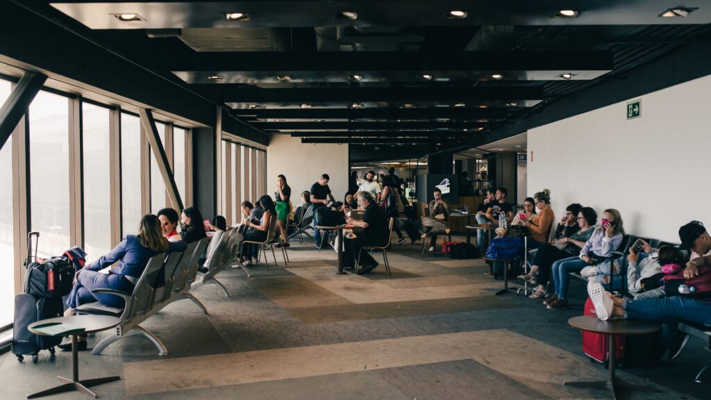 Busy airport waiting area with travelers seated near large windows. Passengers sit with luggage while others use their phones or wait near the gate.
