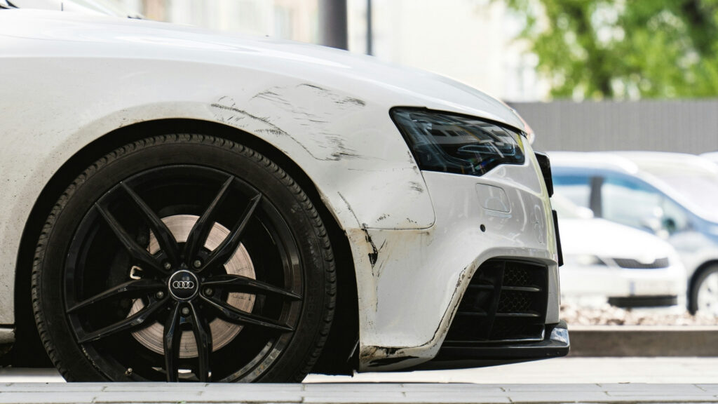 Close view of the front side of a white car with black scrape marks and scuffs across the fender and bumper near the wheel and headlight. The visible damage includes several long scratches and dark paint transfers.
