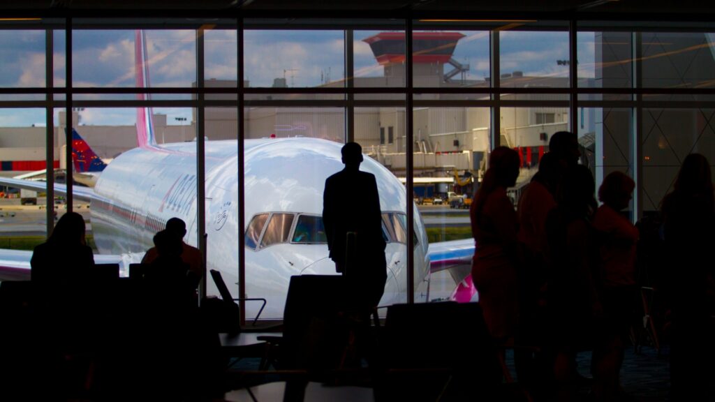 Silhouettes of travelers waiting inside an airport terminal in front of large windows.