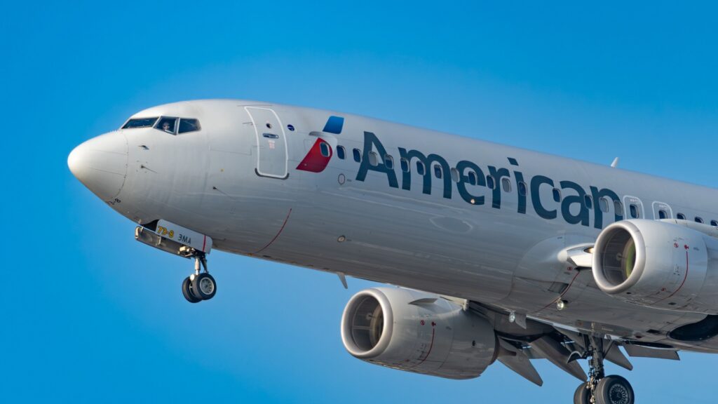 Close view of an American Airlines passenger plane in flight against a clear blue sky. The landing gear is extended and the cockpit nose and two engines are visible.