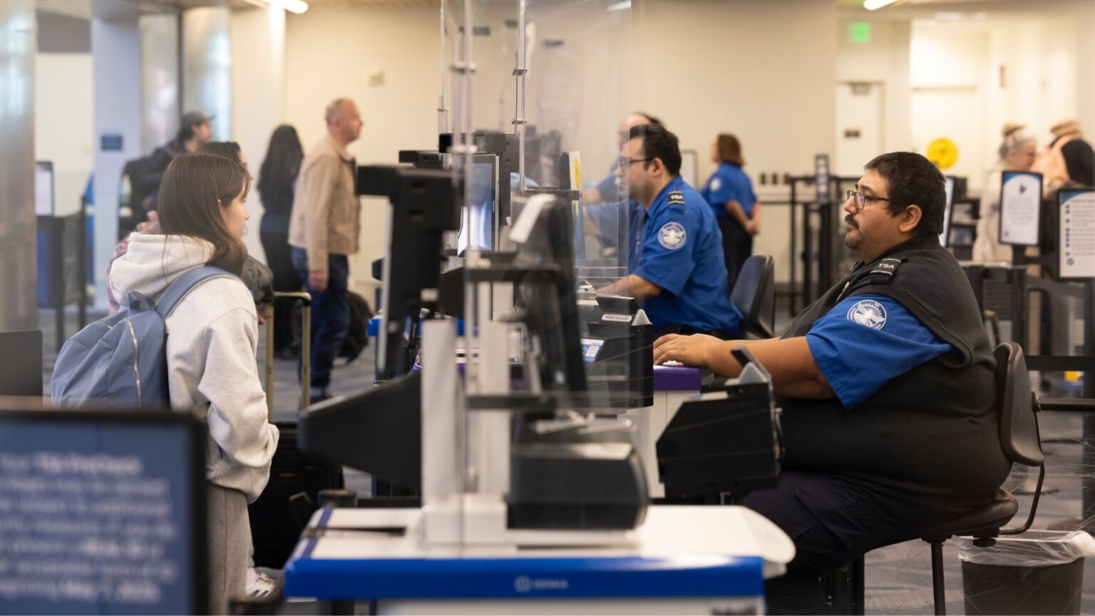TSA Can Fine You $17,000 for Bringing This One Smartphone on a Plane A traveler with a backpack stands at an airport security checkpoint speaking with TSA officers seated behind computer screens and clear partitions. Other travelers and security staff move through the screening area in the background.