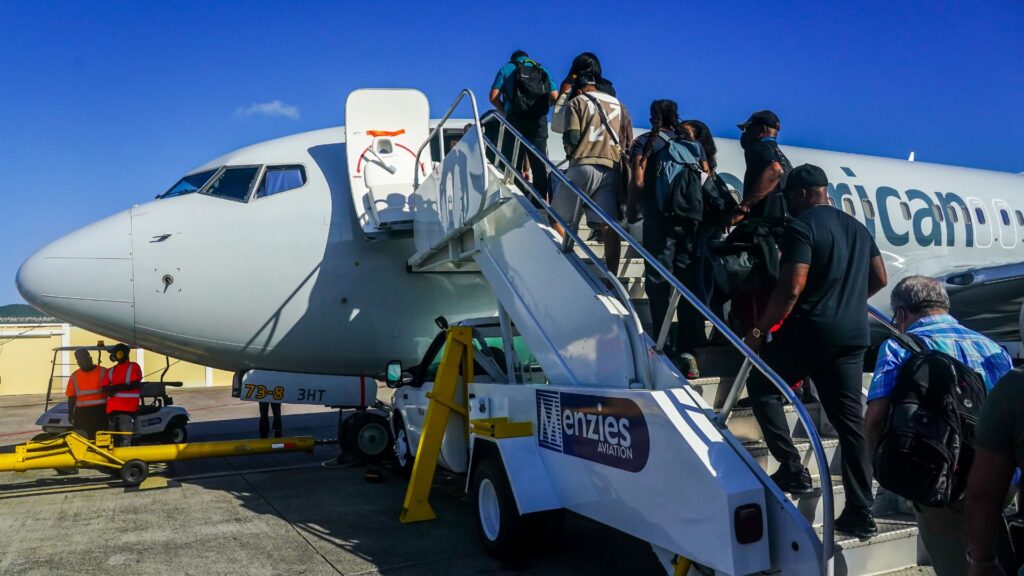 Passengers climb mobile stairs onto a commercial airplane at a sunny airport, carrying luggage.