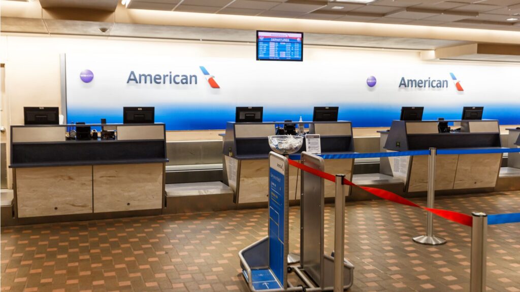 American Airlines check-in counters stand empty behind red barriers, with blue signage, flight screens, and luggage carts.