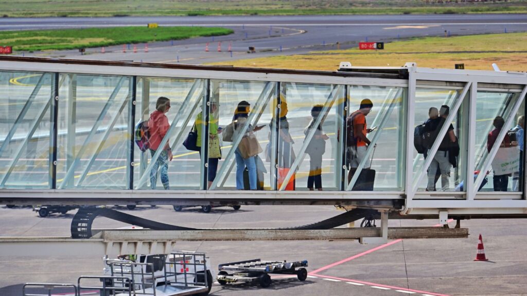 Passengers walking through a glass-walled jet bridge toward a plane, with airport tarmac, baggage carts, and a runway visible in the background.