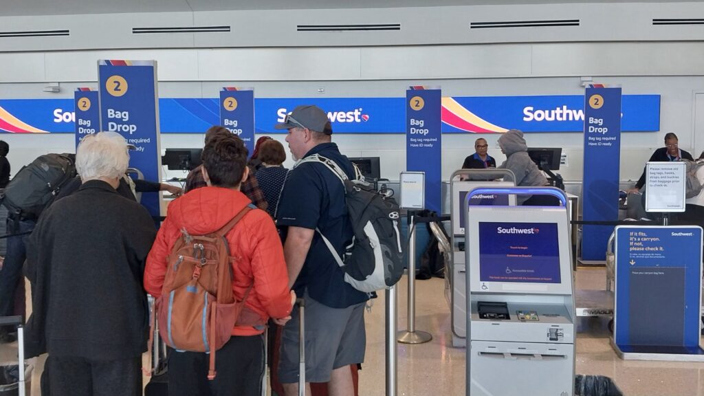 Travelers queue at Southwest Airlines airport check-in counters, with luggage, kiosks, blue signs, and staff visible in a busy terminal.