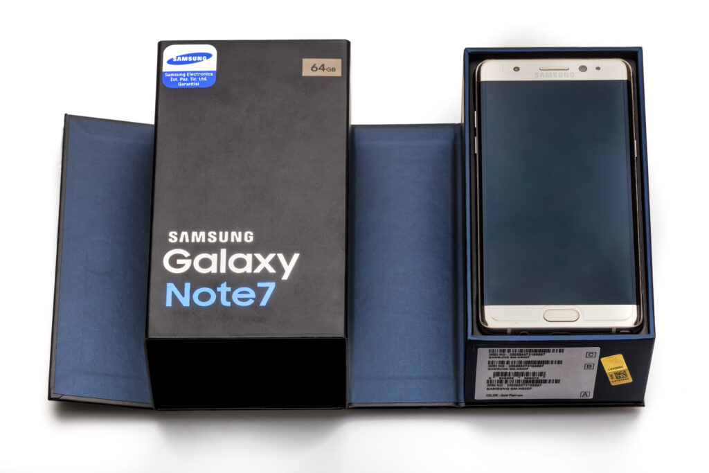 Gold Samsung Galaxy Note7 smartphone rests inside its open dark blue retail box beside a black package labeled Samsung Galaxy Note7 and 64GB. The phone is shown from the front with its curved screen turned off.