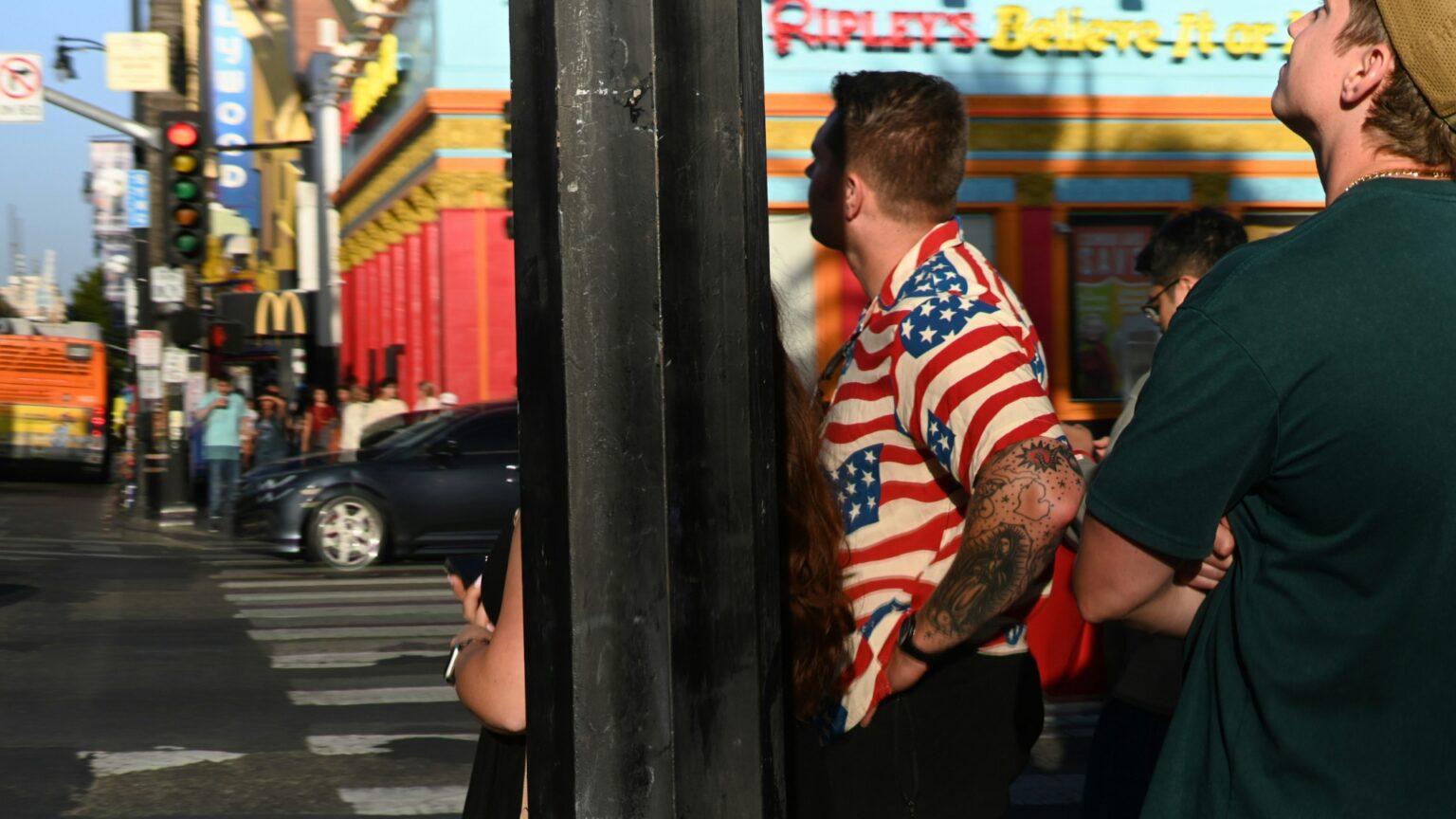 Pedestrians wait at a busy city intersection near a crosswalk. One man wears a shirt patterned with American flags while storefronts and traffic fill the background.