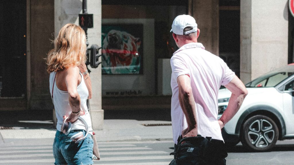 Two pedestrians wait beside a city crosswalk on a sunny day. A woman in a white tank top and denim shorts stands beside a man wearing a pale pink shirt and white cap.
