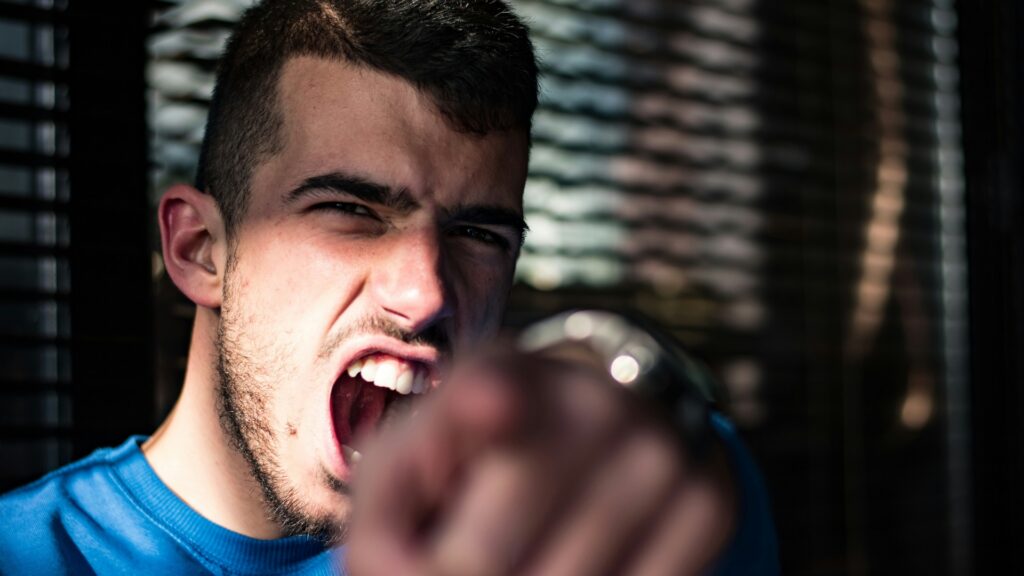 Young man in a blue shirt points directly toward the camera with his mouth open and an intense expression. His outstretched hand is blurred in the foreground.