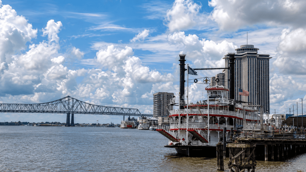 Riverboat docked along the New Orleans waterfront with a bridge and city buildings in the background.