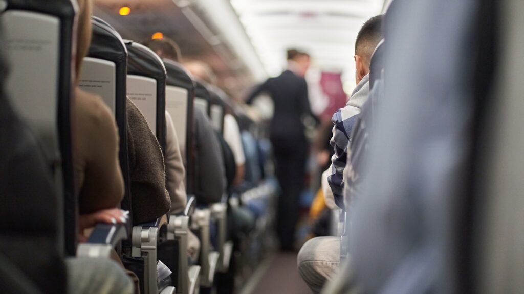 View down the aisle of a passenger airplane with travelers seated on both sides. A person stands farther down the narrow aisle among the rows of seats.