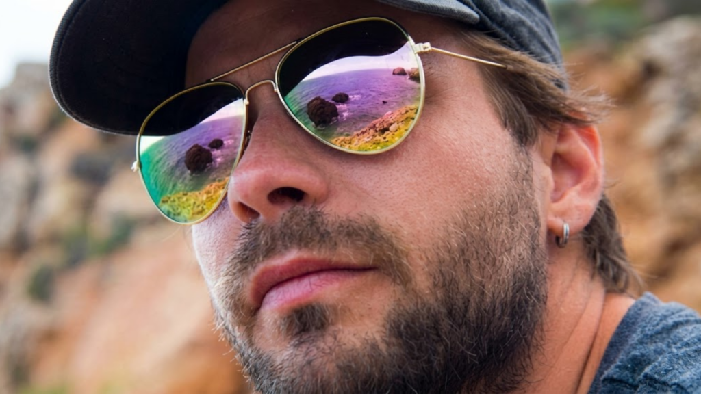 Close-up of a man wearing reflective sunglasses near a rocky coastline.