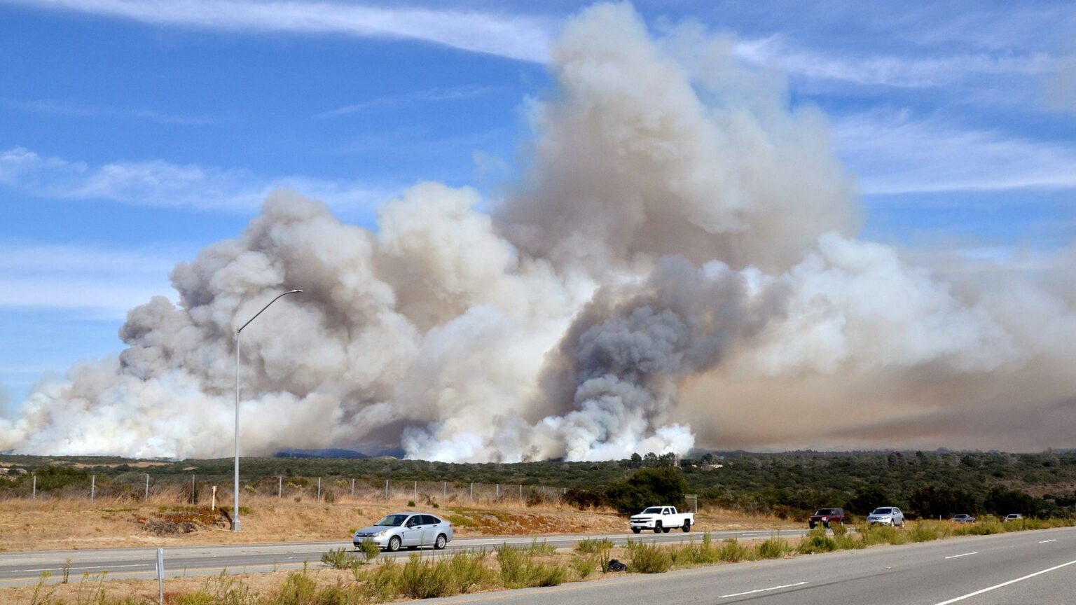 How to Handle Travel During a Raging Wildfire Season Without Ruining Your Trip Vehicles on a rural highway drive past a sudden and fast moving wildland fire.