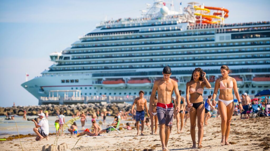 Young guys and girls wearing surf shorts and bikinis walk on the beach as a Carnival cruise ship leaves port in the background.
