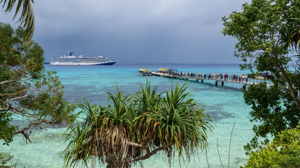 A Carnival Cruise Line luxury liner ship anchored off the coast in the turquoise waters of Santal Bay next to a busy passenger tender dock under a stormy sky.