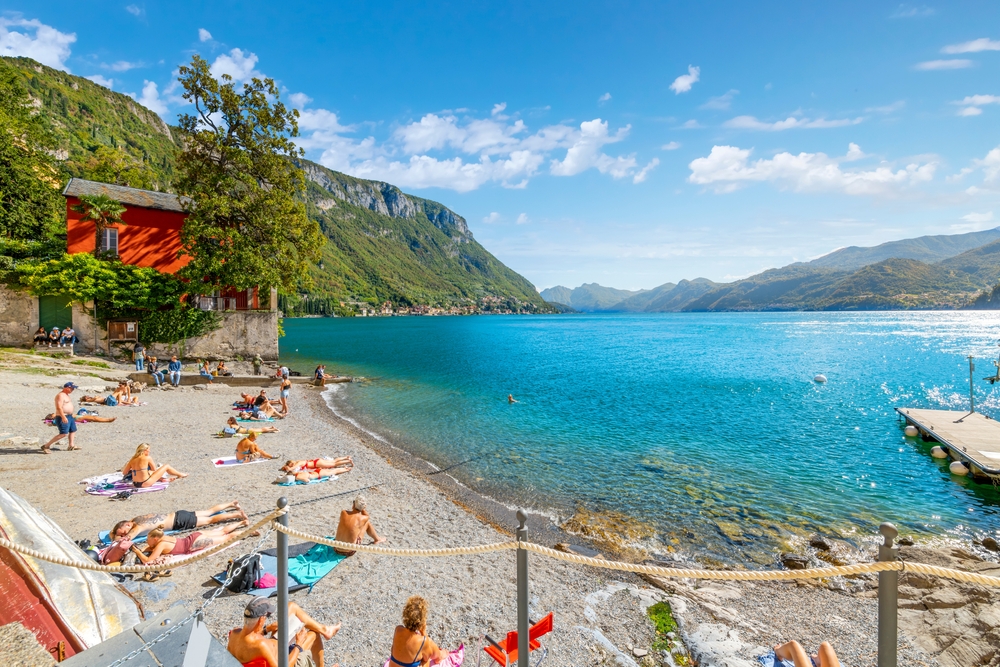 Tourists relaxing on a small beach along Lake Como in Varenna, Italy, with colorful lakeside buildings and mountains in the background.