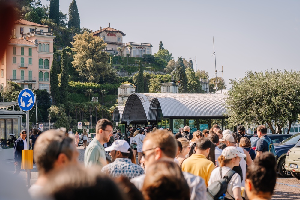 Tourists wait in a long line at the Varenna ferry terminal, with hillside villas and trees overlooking Lake Como.