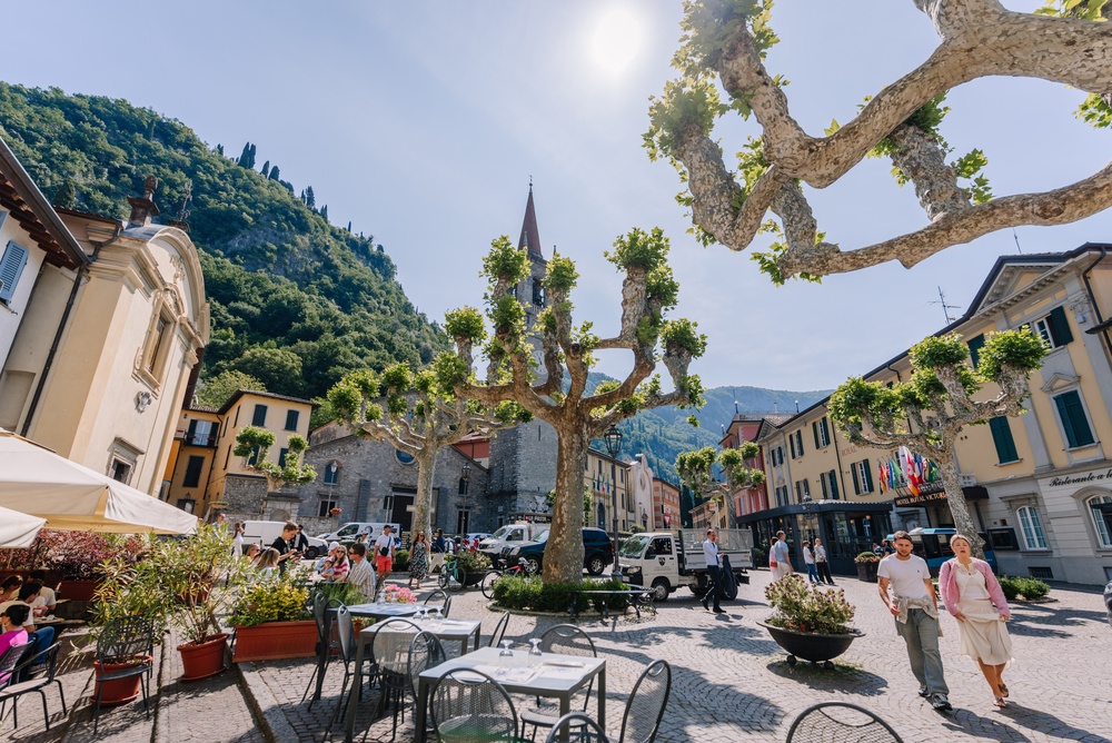 People walk through Varenna's main square, with pruned plane trees, a church bell tower, and surrounding buildings set against a wooded hillside.